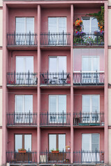 Facade of residential building of pink with balconies, one is decorated with flowers, rest empty with metal railings.