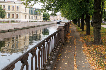 Embankment of water channel with wrought-iron lattice and pedestrian path strewn with yellow autumn leaves. Autumn mood.