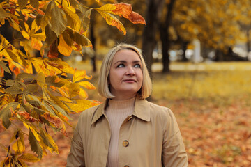 Portrait of a smiling blonde woman in a tan trench coat standing outdoors in an autumn park, with bright orange and gold leaves creating a vibrant fall background. Autumn concept,  autumn season