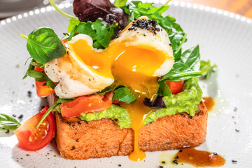 Poached egg on avocado toast with cherry tomatoes, fresh greens, and runny yolk on a white plate