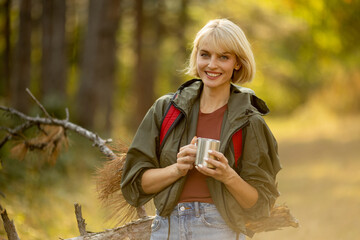 Smiling woman enjoying warm beverage in a sunny forest, wearing a green jacket and backpack during autumn