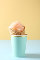 Waffle cone with cotton candy in paper cup on color background, closeup