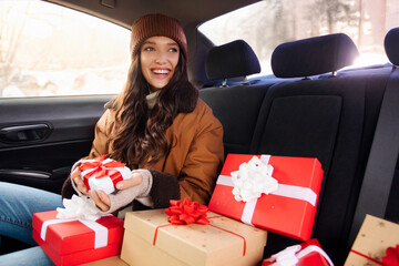 Smiling lady sitting in car on back seat with Christmas gift boxes, going to congratulate friends...