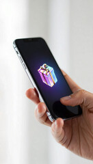 a person's hand holding a modern smartphone, with a glowing holographic gift box floating above the screen