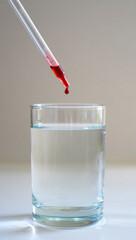 A glass pipette hovers above the glass, with a single drop of red liquid suspended at the tip of the pipette