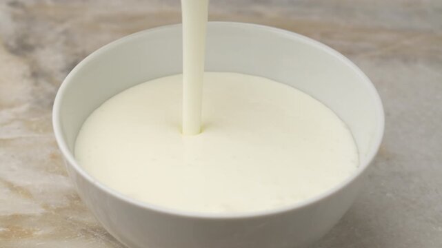 Pouring white dairy product into ceramic bowl on table, close up. Home farm product kefir or milk or yogurt