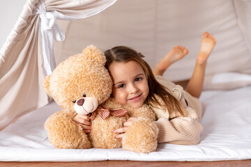 Happy smiling 4-5 years old little girl lying in a wigwam tent, hugging soft toy teddy bear. Children's room, childhood, games, imagination. High quality photo © Irina Mikhailichenko