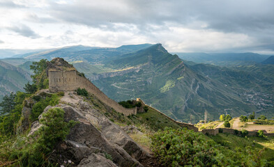 The ruins of a stone fortress sit atop a rocky mountain ridge with green valleys down below in natural daylight