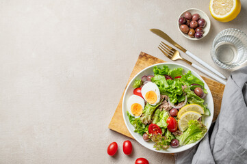 Tuna salad with olives, lettuce, tomatoes, eggs and lemon in a plate on a wooden board on a light background