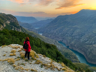 Fototapeta premium A woman in a red jacket stands on a rocky cliff above a lush valley and a winding river at sunset