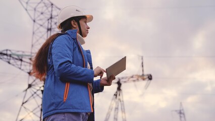 an electrician engineer in helmet works with tablet at sunset in sky, tower under high voltage current, checking electrical volt networks, supplying light to city, industrial safety power engineer.