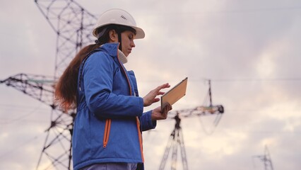 An engineer at a high voltage power plant works in a tablet at sunset in the sky, electrical energy...