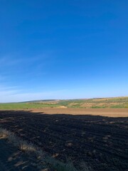 Fototapeta premium Wide rural landscape with burnt soil in the foreground, green fields and hills under a clear blue sky. Concept of agriculture, nature, and ecological contrast.
