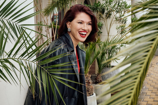 Stylish woman wearing a black coat poses joyfully among lush greenery