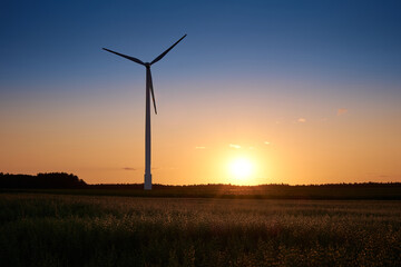 Obraz premium Wind turbine standing in a field at sunset with vivid orange and blue sky, symbol of renewable energy and sustainable future.