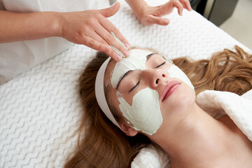 Naklejka premium Cosmetologist applying rejuvenating facial mask on a female face in beauty salon. Facial treatment concept