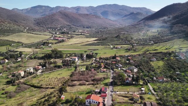 Picturesque aerial shot of Italian countryside village situated in the heart of powerful mountain hills, village with scattered private houses and agricultural fields in sunlight, olive tree groves
