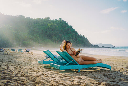 Woman reading book on the beach laying on chaise lounge 