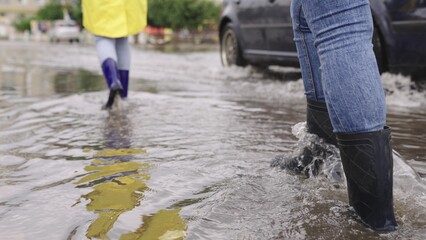 girls in raincoats and rubber boots walk along road flooded with torrential rains, their feet walk...
