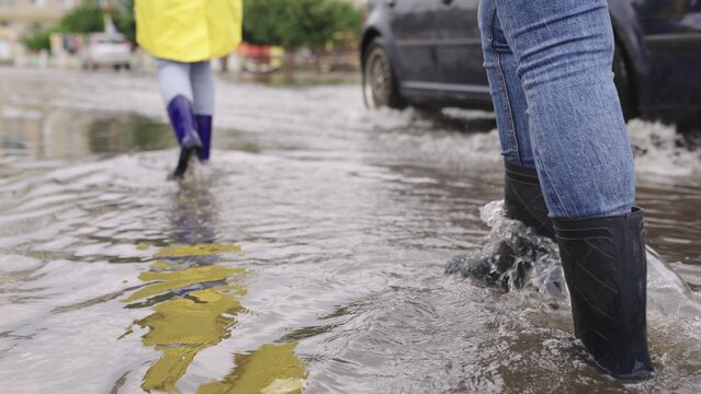 girls in raincoats and rubber boots walk along road flooded with torrential rains, their feet walk through puddles city, splashing water to the sides, the flood is on street, car is driving on water.