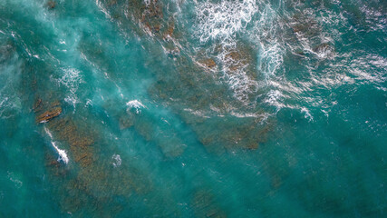 Aerial view of ocean waves breaking over rocks with turquoise blue water and white foam natural sea...