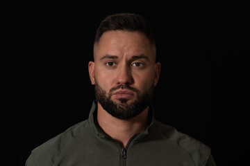 Fototapeta premium Intense studio headshot of a handsome, serious bearded man in military uniform with a confident gaze, isolated on a solid black background.