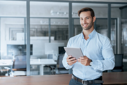 Latin bearded adult professional business man holding digital tablet using fintech tab app standing in company office. Young smiling businessman trader using touchscreen computer. Portrait at camera - Powered by Adobe