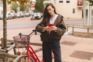 Serious lady standing near bicycle using mobile app outdoors. Busy young woman wear trench coat and jeans, standing near, hold mobile phone, texting message in city street.