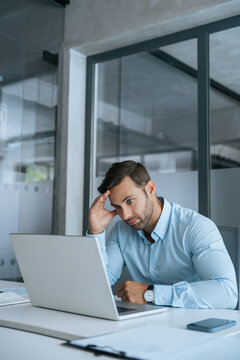Confused young businessman looking disappointed at laptop pc computer screen having stress. European business man holding hand near temples, feeling stressed and tired sitting in office. Vertical