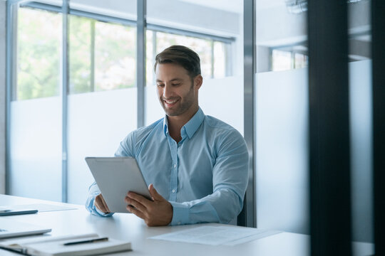 Smiling European entrepreneur businessman looking at digital tablet screen sitting at desk in office. Professional executive man working using computer for financial business banking work. Copy space