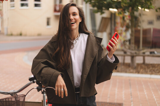 Brunette woman standing near bicycle and hold phone in city, street or urban road outdoors. Bike, travel and happy female using 5g mobile tech, internet browsing or social media, web or online surfing