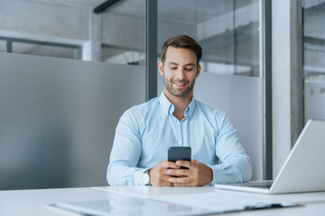 Smiling young Latin businessman holding smartphone telephone device sitting at desk in office. European manager using digital technology cellphone phone mobile, laptop for business work. Copy space