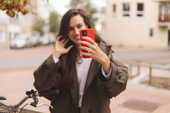 Brunette woman standing near bicycle and hold phone in city, street or urban road outdoors. Bike, travel and happy female using 5g mobile tech, internet browsing or social media, focus on phone.