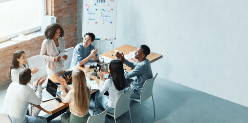 Elegant African American woman holding digital tablet and speaking to colleagues, making business report, high angle view, copy space
