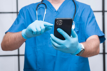 Close-up of a medical professional in blue scrubs and gloves holding a smartphone. Stethoscope around neck. Concept of telemedicine, healthcare technology and online consultation.