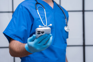Close-up of a medical professional in blue scrubs and gloves holding a smartphone. Stethoscope around neck. Concept of telemedicine, healthcare technology and online consultation.
