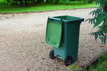 Large green plastic waste bin with wheels and hinged lid positioned on gravel pathway surrounded by lush garden vegetation. Municipal waste management container.