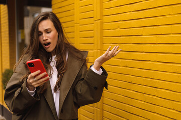 Confused puzzled brunette woman in casual clothes, standing holds a smartphone in her hand, looks...