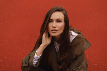 Naklejka premium Portrait of elegant woman sitting outdoor, hold her head, look sensual and dreaming, look at camera. Girl wear raincoat.