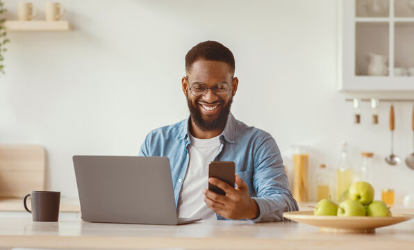 Smiling glad young african american with beard businessman in glasses checks mail on smartphone and works on laptop on minimalist kitchen interior. Video blog, modern technology and social distance