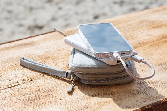 Mobile phone with clear protective case connected to white power bank placed on gray zippered carrying case on wooden surface. Travel electronics kit for mobile device backup power.