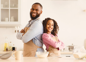 Home Bakery Concept. Portrait of happy african american dad and his child daughter standing back to back in kitchen and posing together, man and girl wearing aprons and making pastry