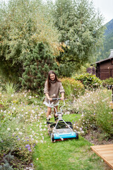Smiling young woman mowing lawn with manual push mower in colorful garden in chalet. Sustainable living and gardening in mountains. Countryside outdoor leisure and lifestyle