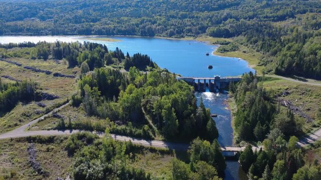Panoramic aerial view of the old Mitis Dam, which controls the water level at the outlet of Lake Mitis, surrounded by forest, woodsheds, and logging roads. Quebec, Canada, 2025.