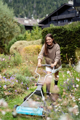 Smiling young woman mowing lawn with manual push mower in colorful garden in chalet. Sustainable living and gardening in mountains. Countryside outdoor leisure and lifestyle