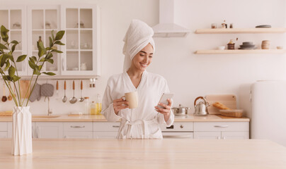 News in app, fresh, good morning and delicious coffee at home. Smiling beautiful young european lady in robe, towel on head drinks and typing on smartphone in minimalist modern kitchen interior