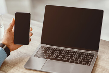 Millennial african american guy working on laptop, hold smartphone with empty screen in office home interior at workplace, cropped. Technology for successful business, app, advertising and website