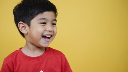 Little boy with short black hair smiling widely, showing his teeth. He wearing red t-shirt, standing against yellow background. His eyes closed he laughs, capturing moment of pure joy and happiness