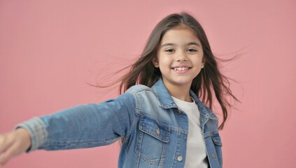 Smiling young girl with long brown hair, wearing denim jacket over white t-shirt, poses against pink background with her arm extended