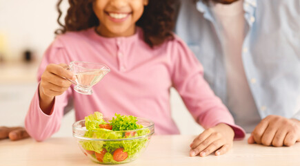 Healthy Food. Close up portrait of smiling black girl adding and pouring olive oil in glass bowl with fresh vegetable salad, cooking together with father at home, selective focus, blurred background